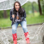 Happy girl jumping in puddles in rainboots, laughing, holding an umbrella above her.